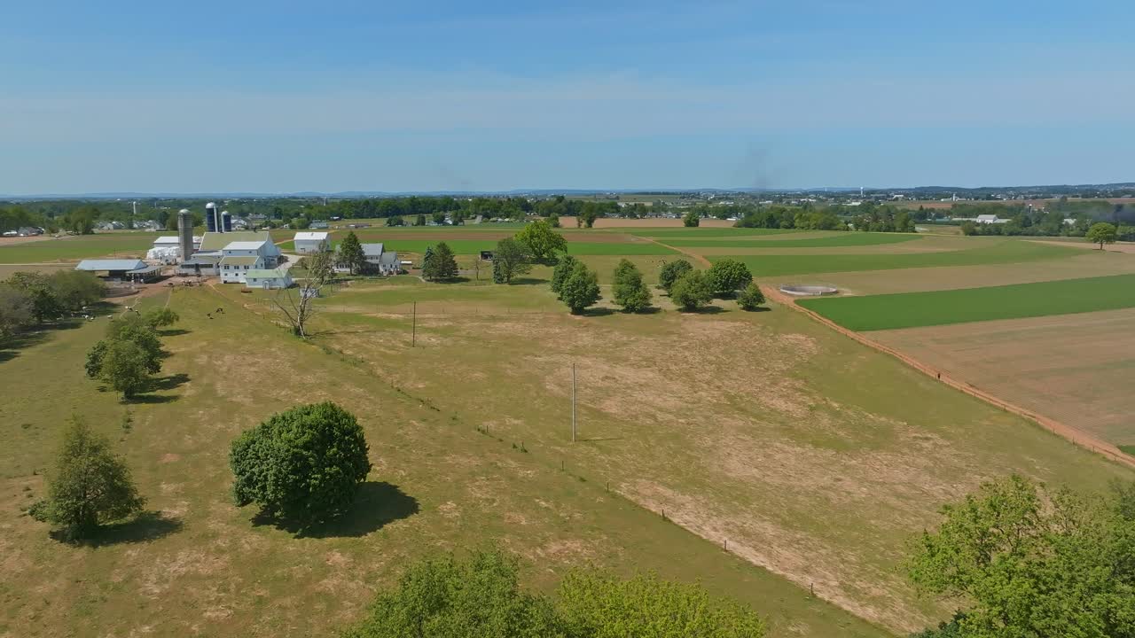 A vast landscape showcases green fields and a serene farmhouse surrounded by lush trees. The bright blue sky complements the rural beauty, creating a peaceful atmosphere of nature.