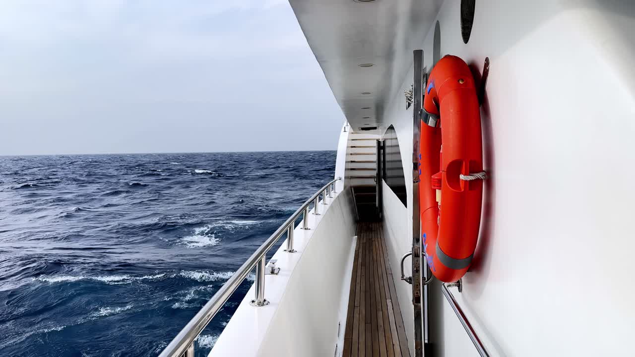Malerisches Seitendeck einer weißen Luxusyacht, die an einem hellen, sonnigen Sommertag durch tiefblaue, kabbelige Mittelmeerwellen fährt, mit einem leuchtend orangefarbenen Rettungsring an der Seitenwand und Holzboden.