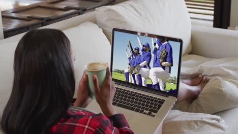 composición de una mujer sentada en casa sosteniendo café viendo un juego de béisbol durante la covid 19 en la televisión