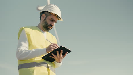 on a sunny day, a professional caucasian engineer wears a white helmet and reflective vest while using technology to audit wind turbines in a field of renewable energy generators