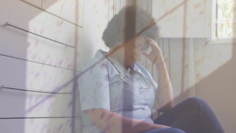 animation of rain on window over unhappy african american female doctor sitting in corridor