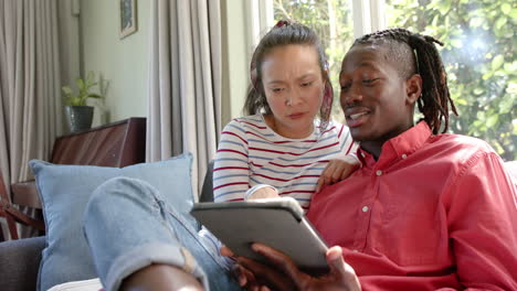 happy diverse couple sitting on couch using tablet in sunny living room, slow motion