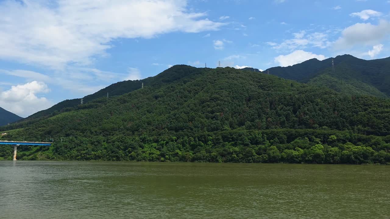 Wide landscape showing Sincheongpyeongdae Bridge crossing Bukhangang River with Hwayasan Mountain in lush green forests under bright summer sky