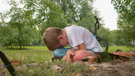 un niño está sentado en una alfombra al aire libre, con la cabeza inclinada cerca de un tronco de árbol, una bicicleta verde yace en el suelo, y una bicicleta negra está estacionada detrás en un campo abierto