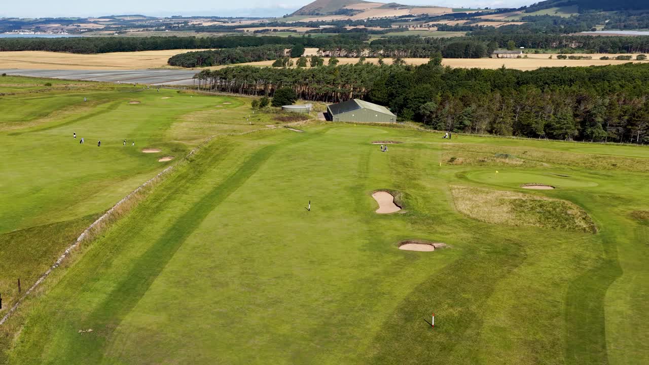 Drone glides above green fairway, sand bunkers, and clubhouse under bright daylight in Scotland