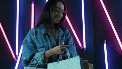 young girl in blue denim jacket with shopping bags on neon lamps background.