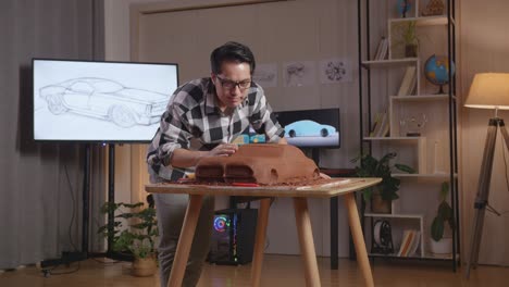 un diseñador automotriz asiático revisa su escultura de arcilla de automóvil después de usar una espátula para suavizar la superficie y crear detalles en el diseño en el estudio