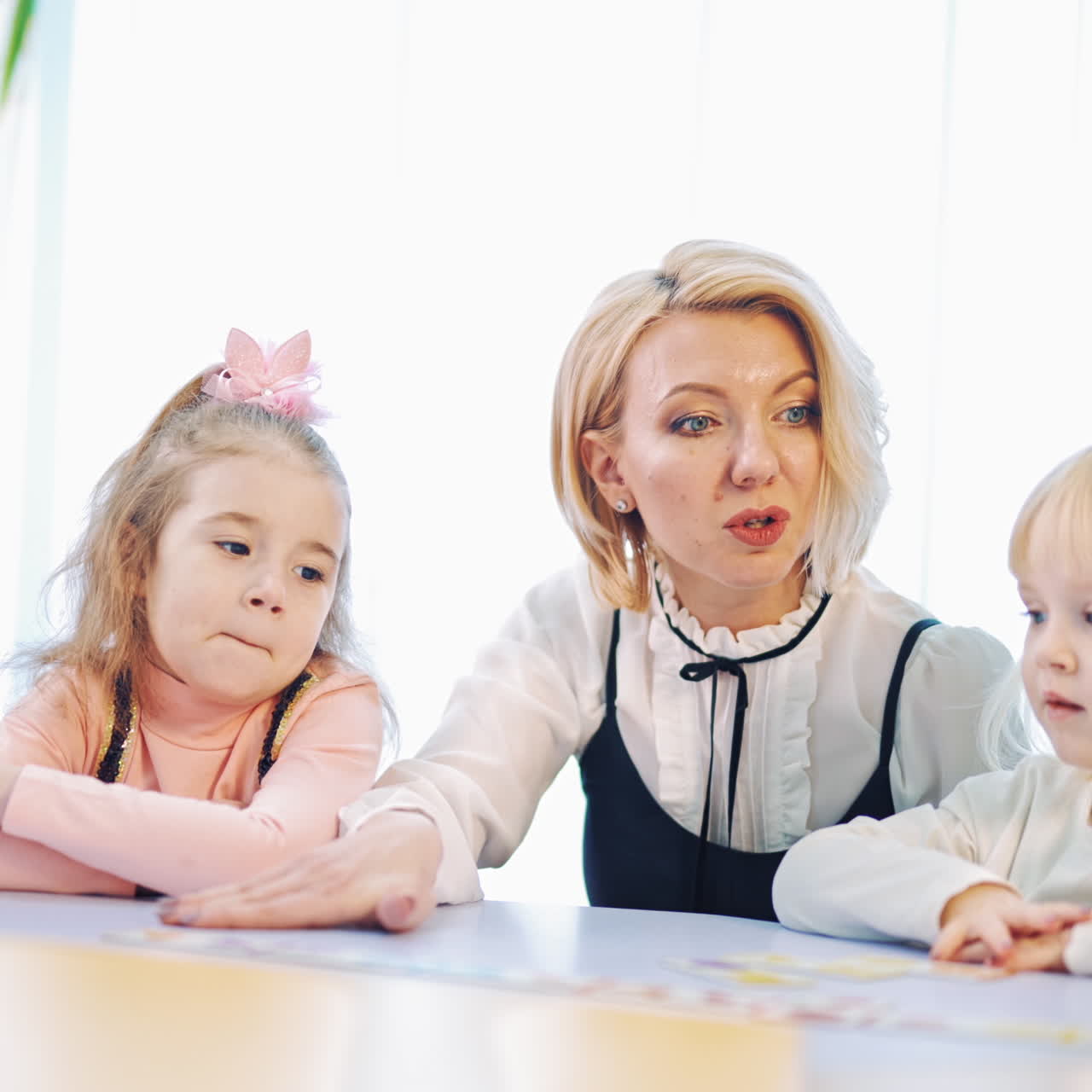 Teacher teaching sitting close to little kids. Small girls listen to their teacher in the classroom. Woman with children at the table.