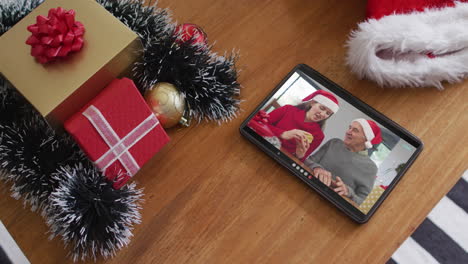 padre caucásico sonriente con su hija usando sombreros de santa en una videollamada de navidad en una tableta