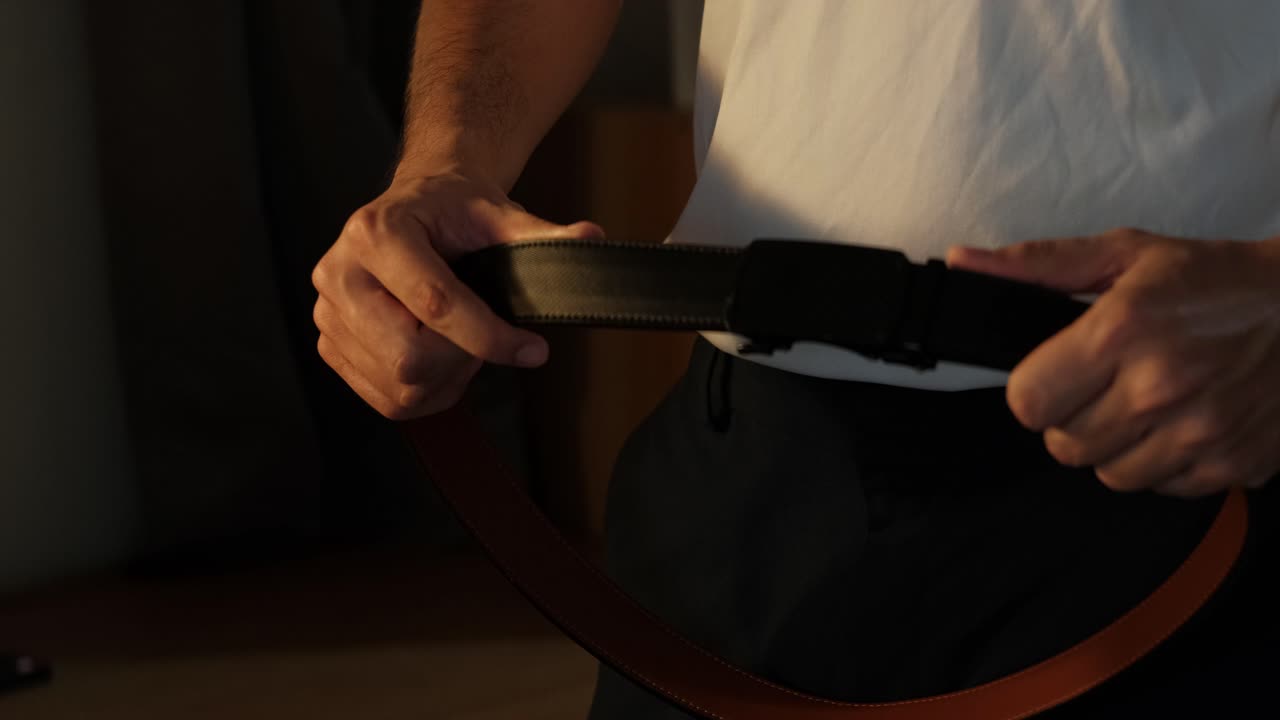 Pengambilan gambar statis seorang pria muda membuka dan menyesuaikan sabuk ratchet kulit untuk tujuan demonstrasi, fesyen gaya hidup