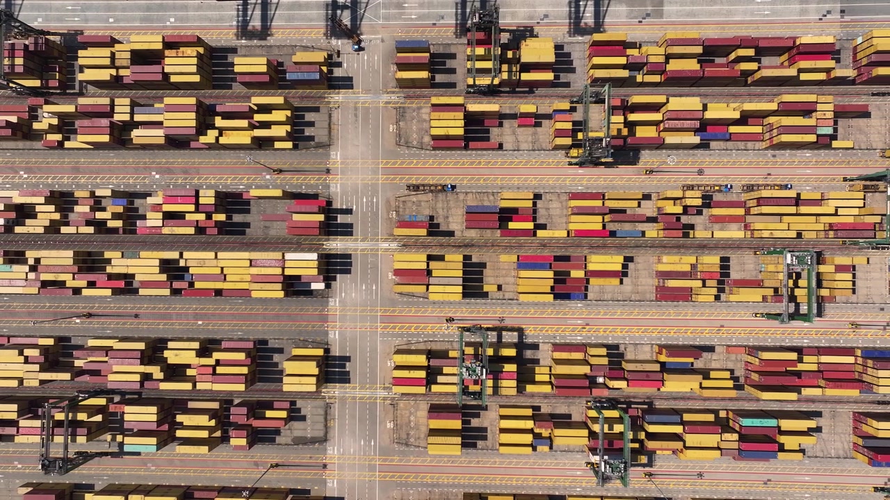 Containers are arranged in stacks at a busy port. Equipment is moving containers. Trucks are loading goods for transportation. Operations highlight the complexity of international trade.