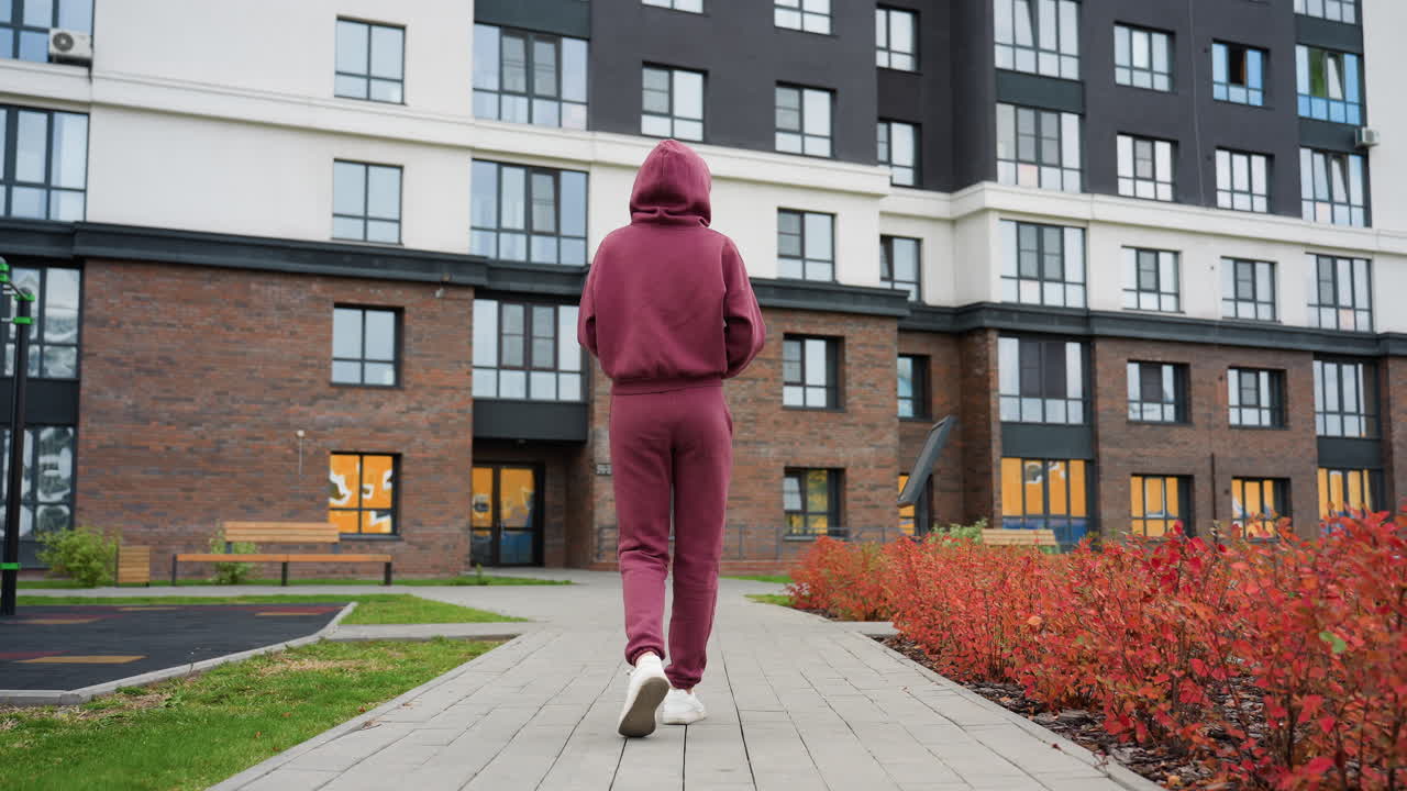 Back view of calm focused woman in maroon hoodie holding water bottle walking toward modern gym building past spring flowers and budding shrubs on paved path in urban training environment