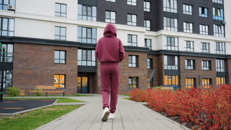 back view of calm focused woman in maroon hoodie holding water bottle walking toward modern gym building past spring flowers and budding shrubs on paved path in urban training environment
