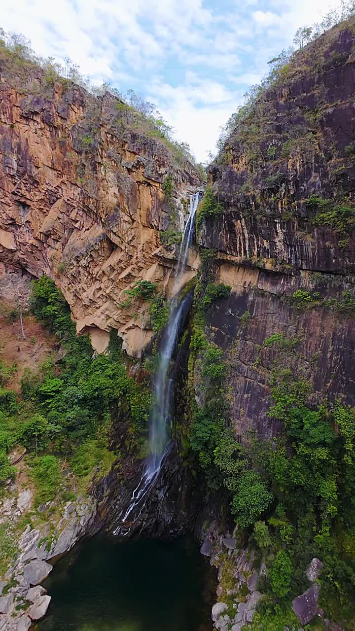 Lush green vegetation lining the tall waterfall cascading down rugged Catua cliffs into a deep natural pool in Chapada dos Veadeiros, Cerrado, Brazil, drone ascending and push in, vertical fpv shot
