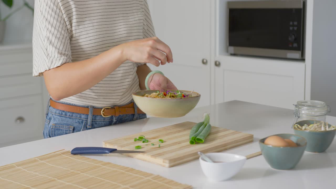 Mujer afroamericana sosteniendo un cuenco, esparciendo cebolla en la isla de la cocina con un cuchillo, terminando de emplatar.