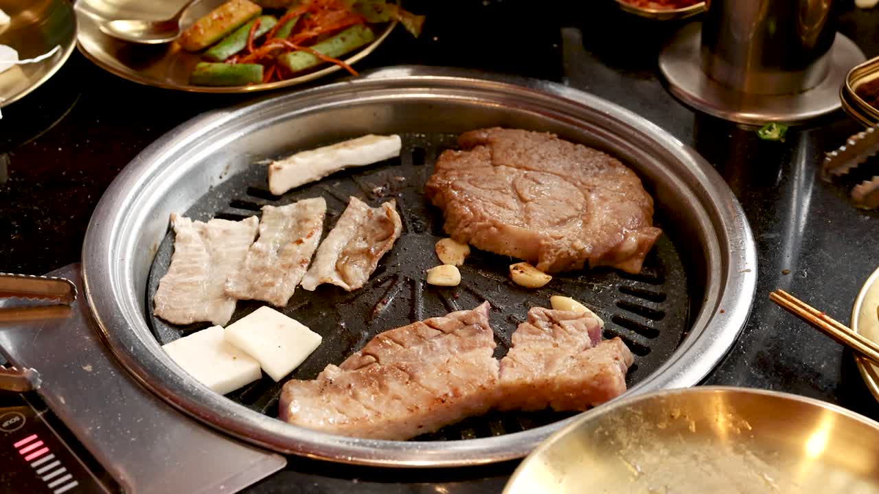 Hand uses tongs to flip pork and garlic on tabletop Korean barbecue grill, warm lighting