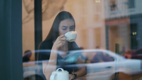 view from the street of a young woman drinking tea or coffee from the white cup in cafe sitting in the coffee shot by the window