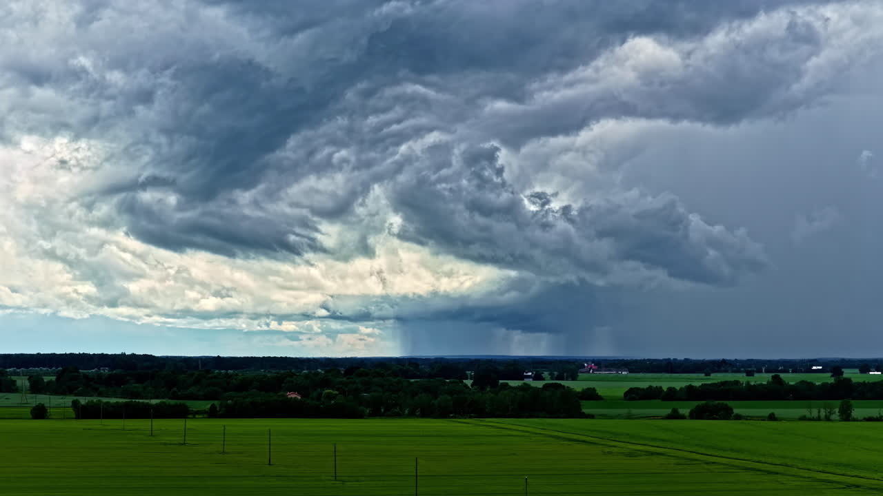 A descending drone captures an ominous and dramatic view of turbulent storm clouds and a heavy downpour of rain approaching a vast green crop field in the Latvian countryside before a thunderstorm