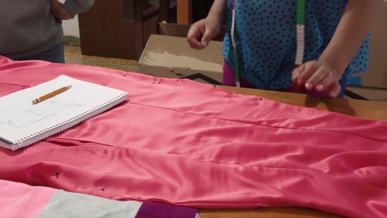 노트북을 사용하여 메모를 작성하는 나무 테이블에 분홍색 드레스를 측정하는 라틴 패션 디자이너