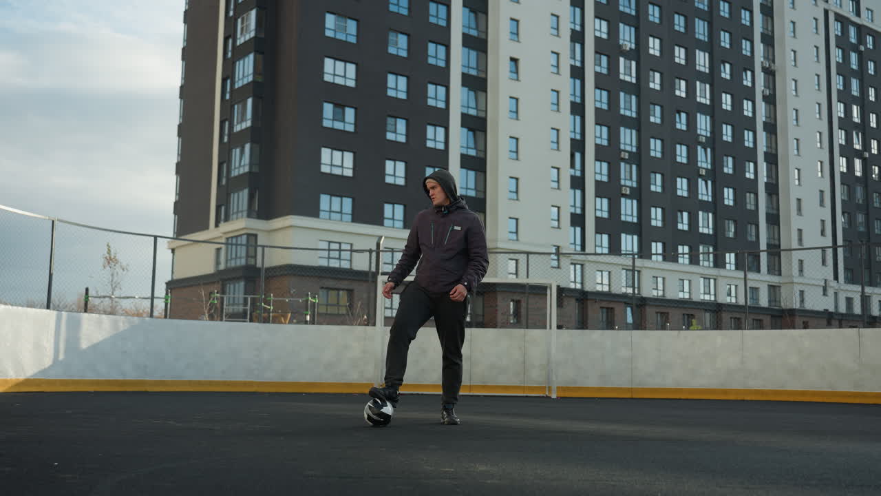 Coach stands in urban sports arena with one leg resting on soccer ball, turning head and arm in a reflective pose, surrounded by towering residential buildings under overcast sky