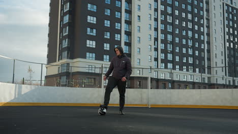 coach stands in urban sports arena with one leg resting on soccer ball, turning head and arm in a reflective pose, surrounded by towering residential buildings under overcast sky