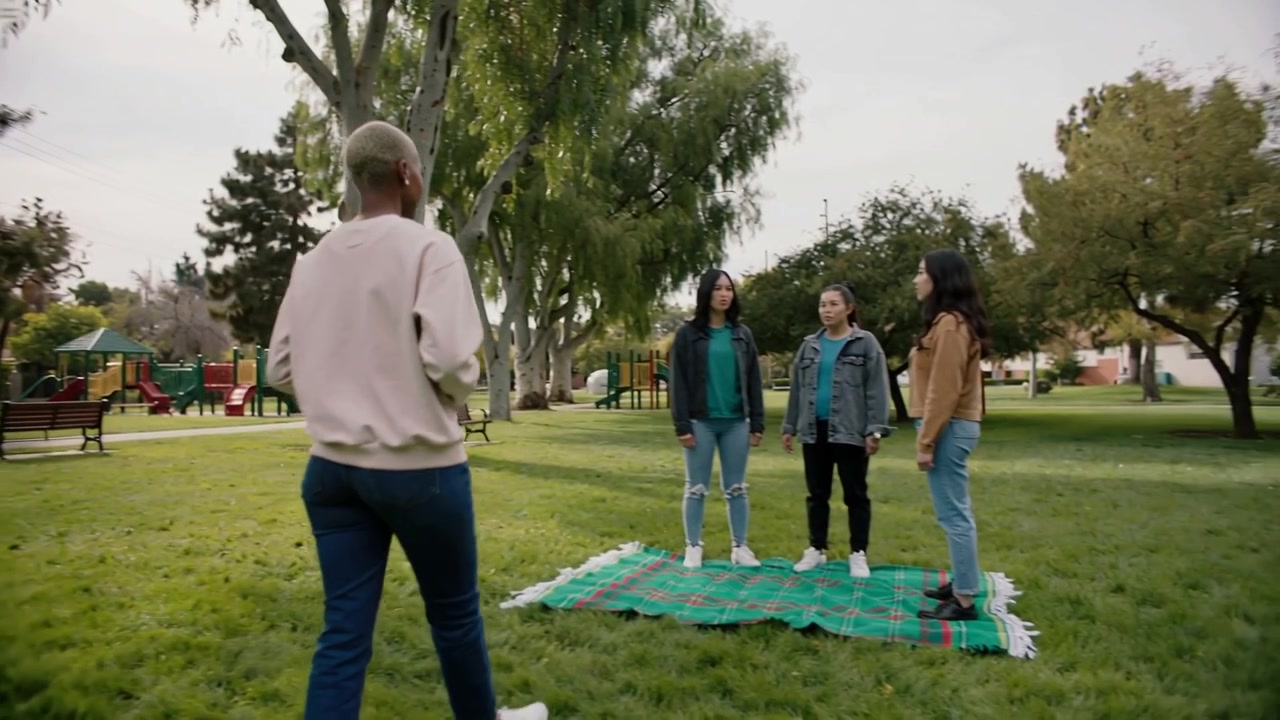 Adulto caminando con una parte de arriba clara, llevando un termo negro hacia una manta de picnic a cuadros, dirigiéndose a encontrarse con adolescentes.