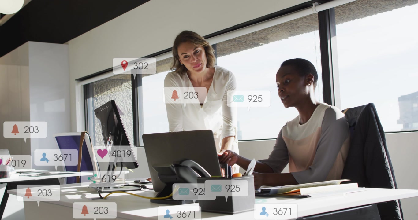 Seated colleague clicking, standing colleague pointing, social icons rising around laptop for ads