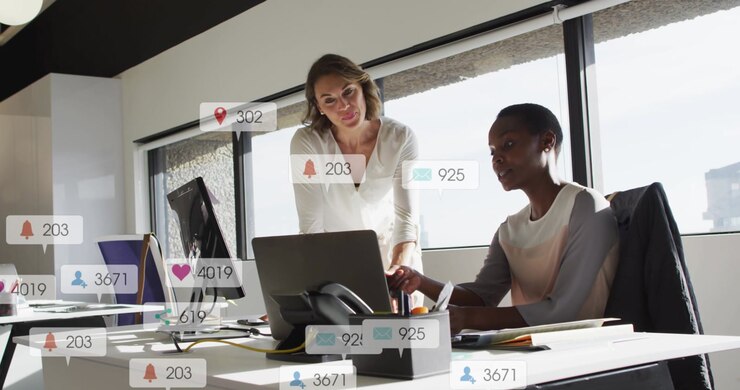 Seated colleague clicking, standing colleague pointing, social icons rising around laptop for ads