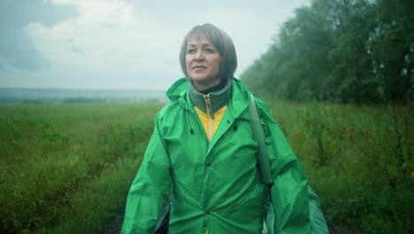 woman in green raincoat carrying mint-colored bag, walking along muddy path surrounded by greenery and trees under misty sky, looking contemplatively into distance