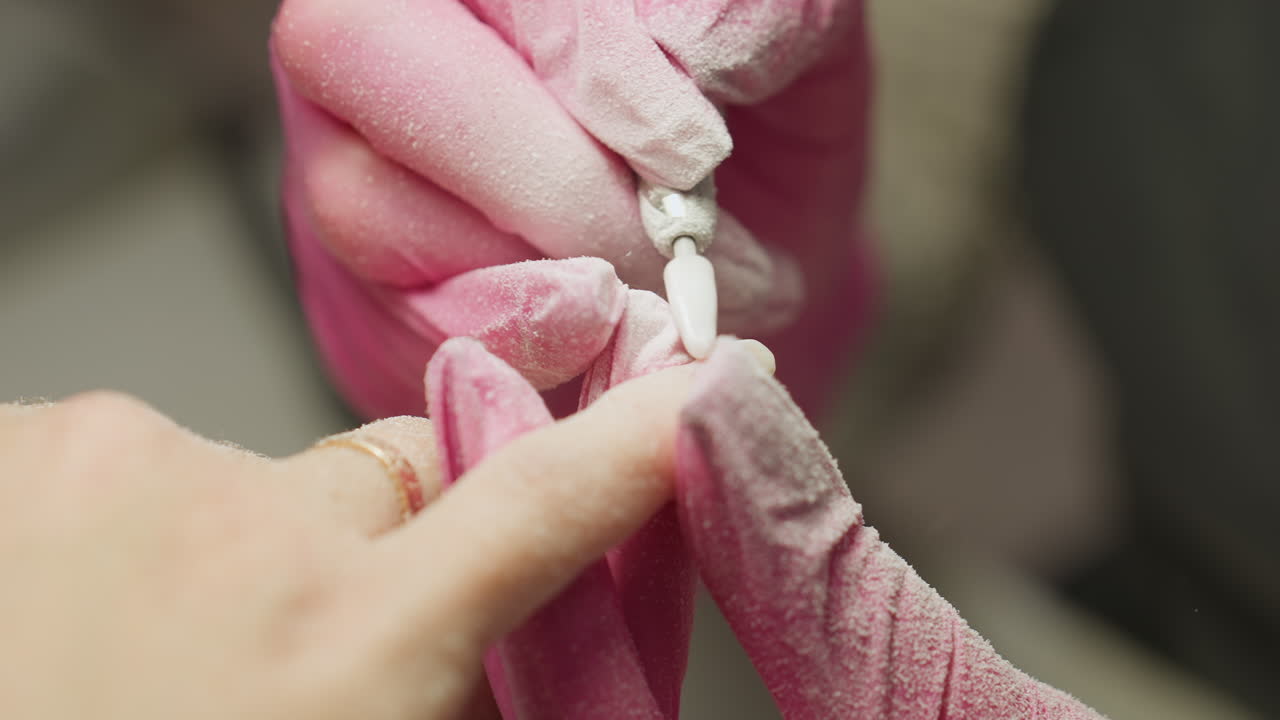 Close-up view of nail technician in pink gloves carefully filing client fingernail using electric nail drill. Client wears gold ring, and filing dust is visible on gloves during precision nail shaping