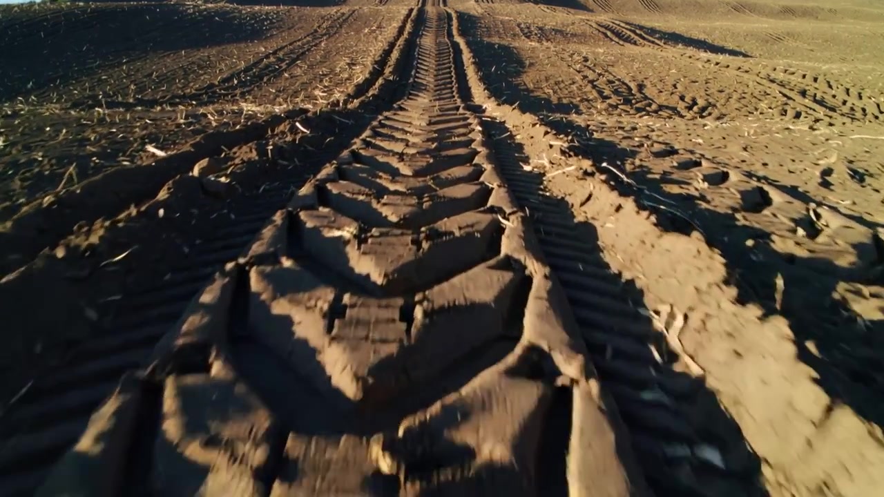 Telecamera in movimento che segue il battistrada di un pneumatico di trattore verso l'orizzonte su un campo, mostrando i tasselli e le zolle.