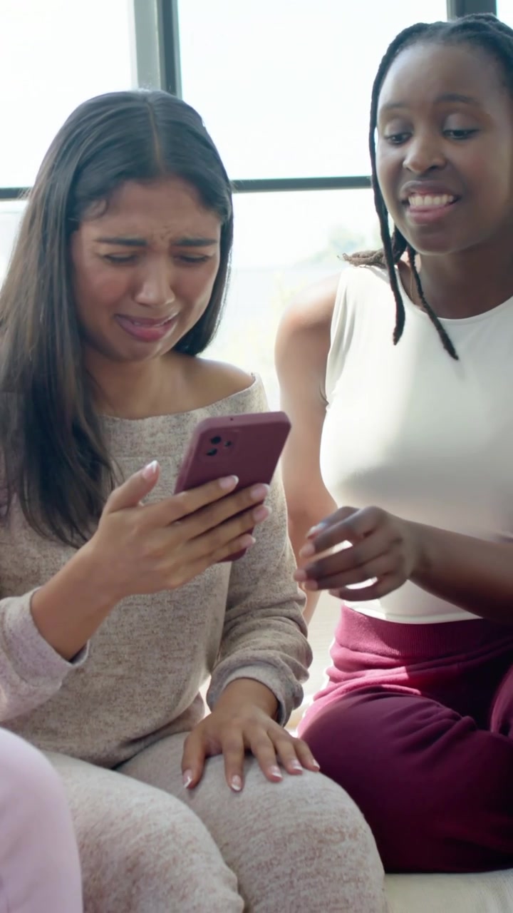 Vertical video: Smiling woman in pants holding trinket, reacting to friend showing phone on sofa