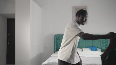 man in white t-shirt carrying backpack, walking in minimalist bedroom, with green headboard, neatly made bed, and window in background, preparing to leave home in bright, modern setting