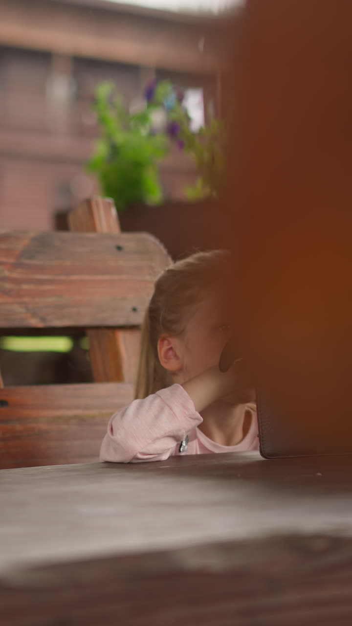 niña pequeña juega videojuego en línea en una tableta sentada en una gran mesa de madera en un café al aire libre en un eco resort en un día de lluvia en cámara lenta