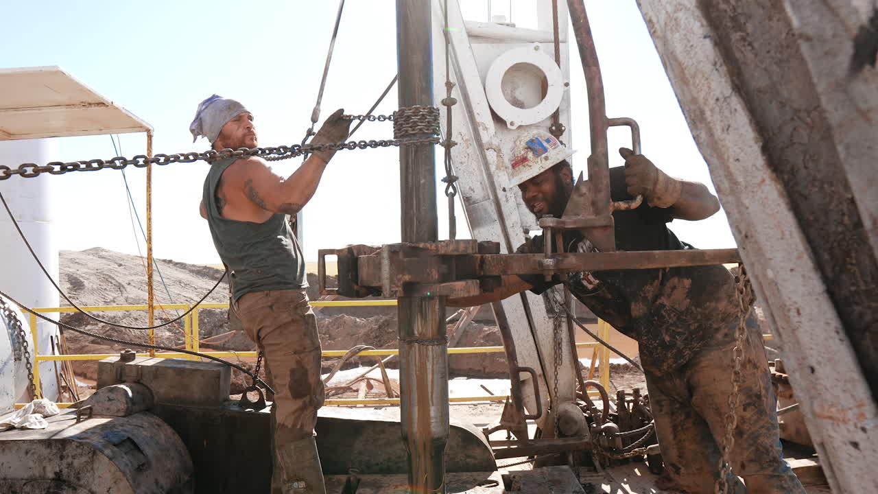 Strong bearded man is holding a chain winded around the metal rotating pipe. Afro-American worker pushes the equipment on the same pipe. Men working at the site for oil production.