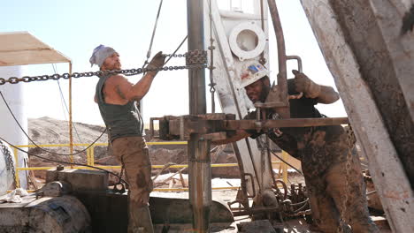 strong bearded man is holding a chain winded around the metal rotating pipe. afro-american worker pushes the equipment on the same pipe. men working at the site for oil production.