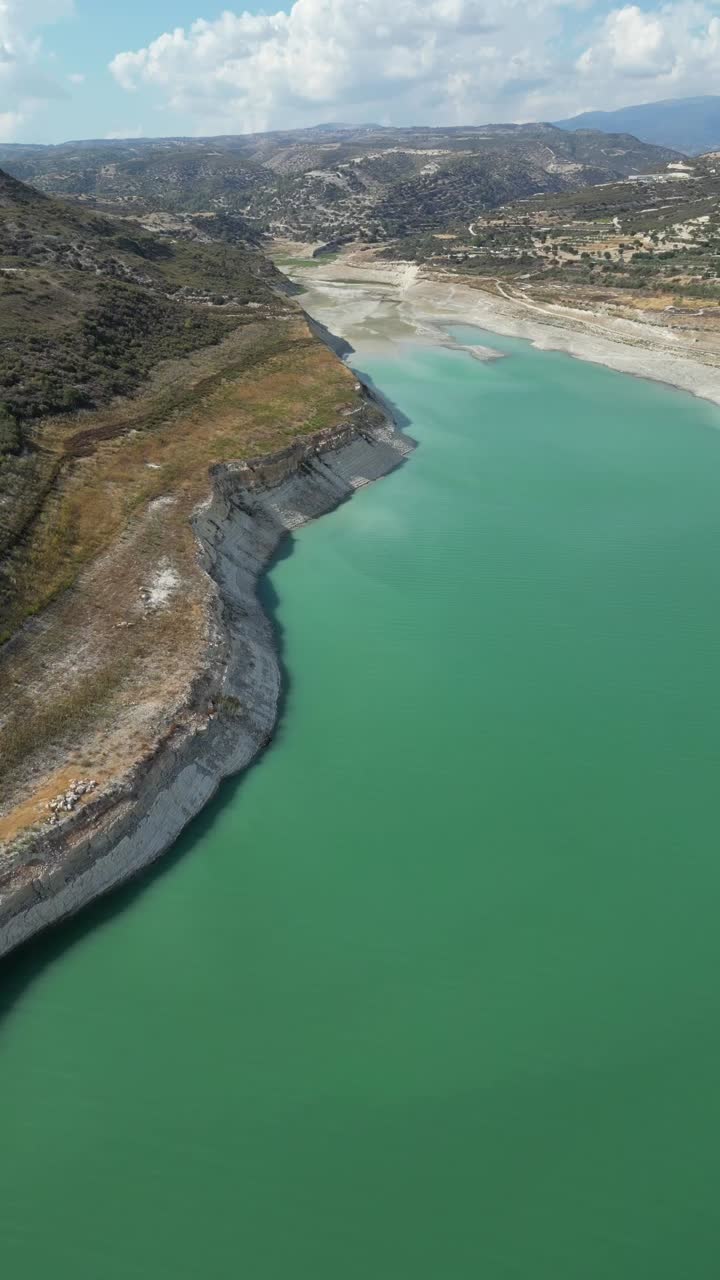 A vertical drone capture reveals the grandeur of Kouris Dam, emphasizing its vast reservoir, winding edges, and the surrounding rugged terrain in stunning detail.