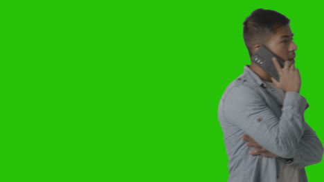 studio shot of casually dressed young man walking across frame talking on mobile phone against green screen