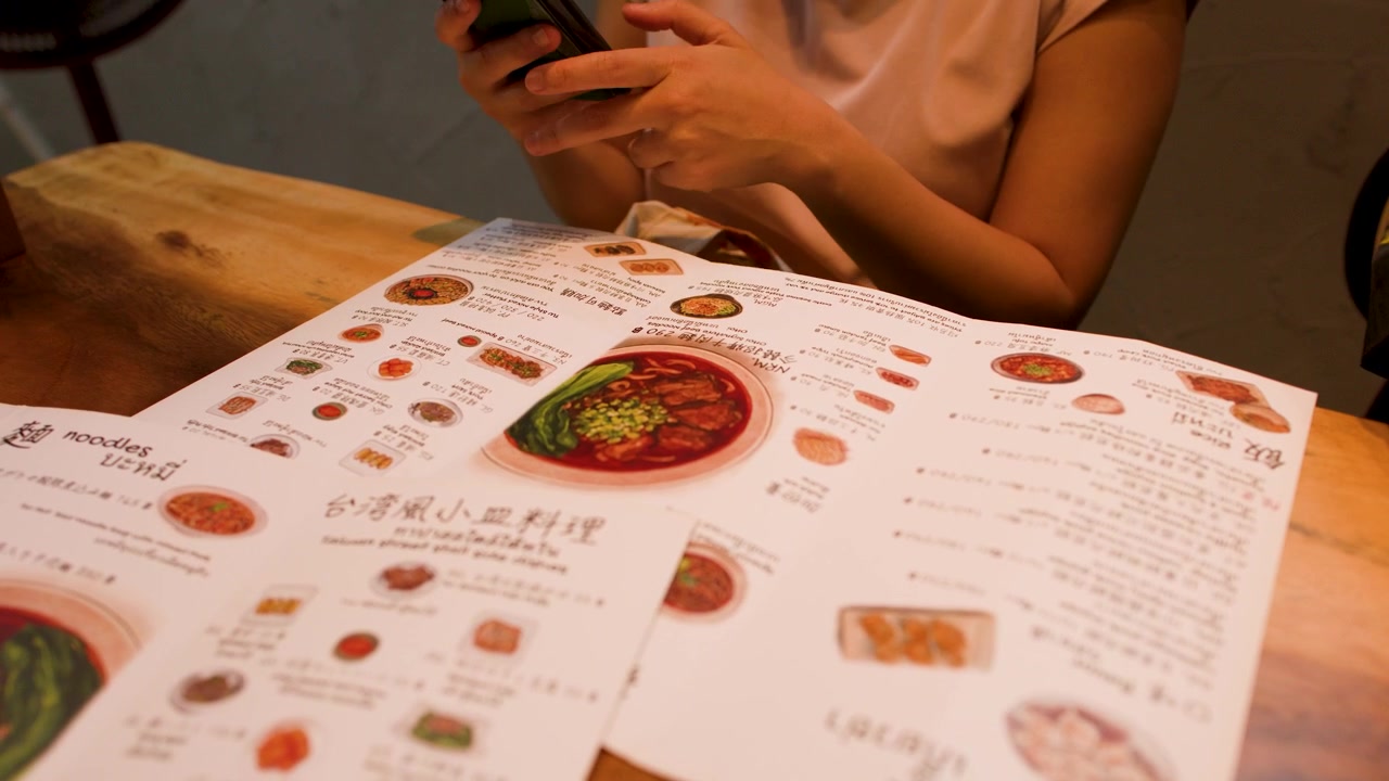 Woman holding a smartphone while browsing a restaurant menu at a rustic wooden table.
