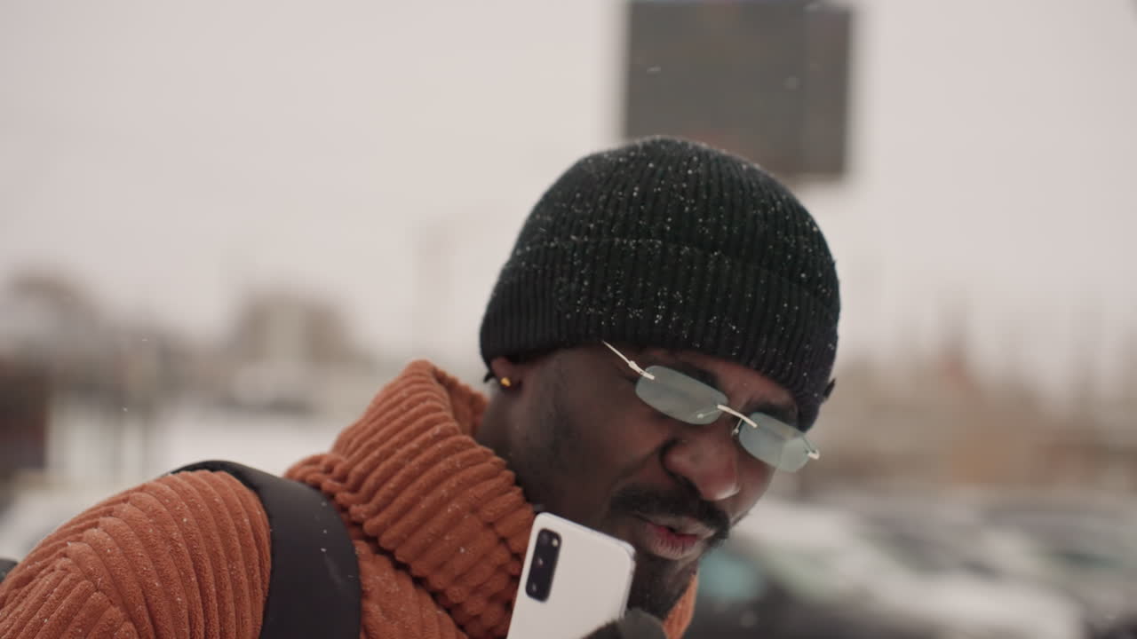 Uomo al telefono in una strada urbana innevata, primo piano di una conversazione con maglione a coste e berretto. Una mano guantata tiene lo smartphone, leggere raffiche di neve. Sullo sfondo un parcheggio, un momento spontaneo di pendolarismo invernale.