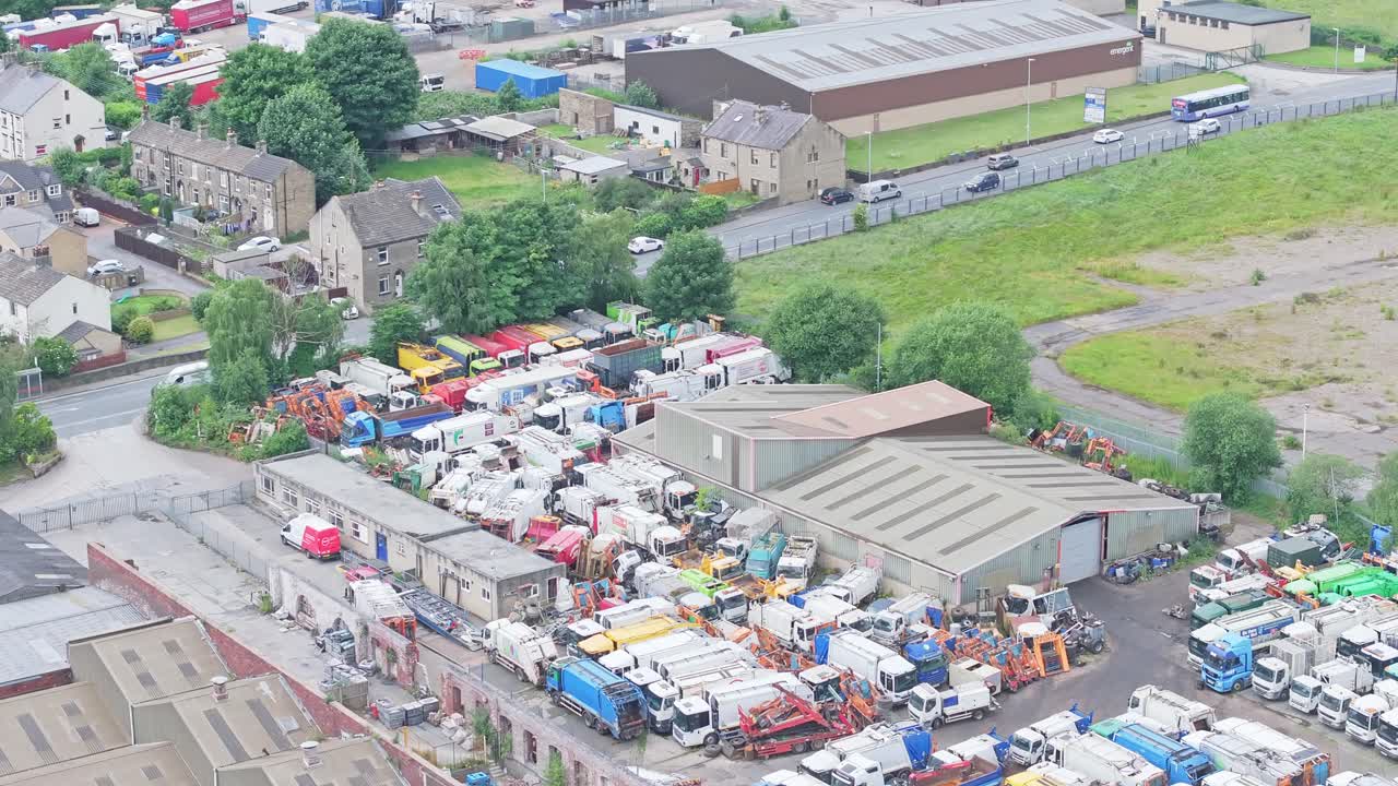 An aerial shot of a busy scrapyard with a variety of dismantled vehicles and machinery parts scattered across the site near industrial buildings. The cluttered area contrasts with the greenery