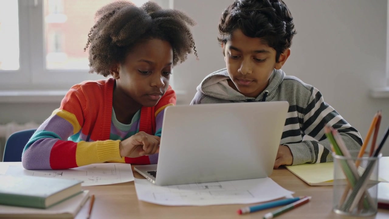 Tapping girl in striped sweater guiding boy in gray hoodie at desk for schoolwork, using laptop