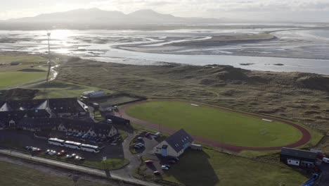 Wide-angle-drone-shot-circumnavigating-the-village-of-Linaclate-in-Benbecula,-featuring-the-surrounding-coastline,-a-wind-turbine,-a-track-and-football-pitch,-distant-mountains-and-the-local-beach