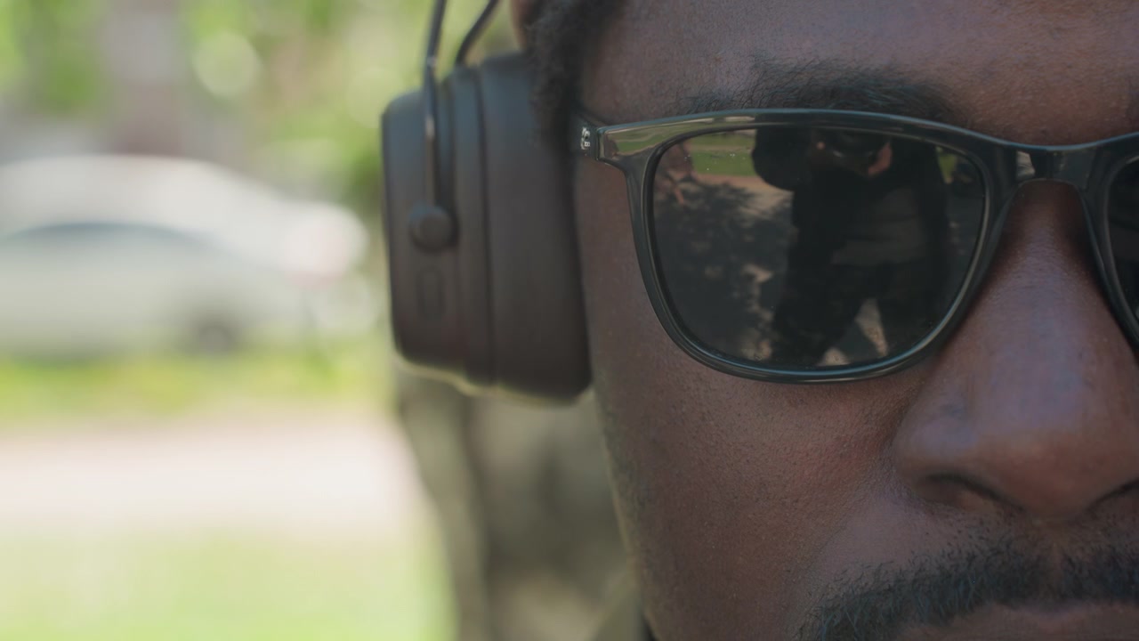 Close up of man wearing black sunglasses and brown corduroy cap with large over ear headphones. Reflections in glasses show another man holding camera outdoors on green grass background