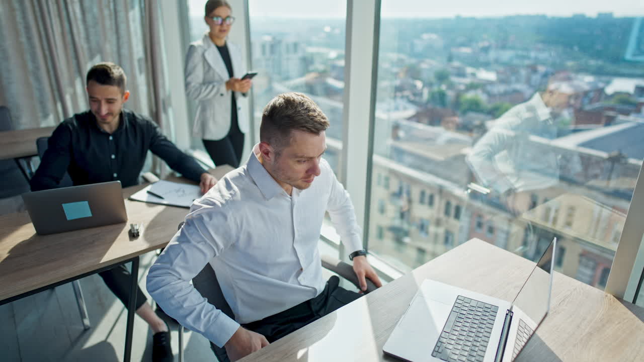 Men in the office working on computers. Blonde lady with phone in hand comes up to a man in white shirt talking to him and smiling.