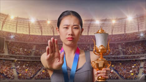 un primer plano de una mujer asiática con una medalla de oro y un trofeo desaprobando sin signos de mano y sonriendo a la cámara en el estadio