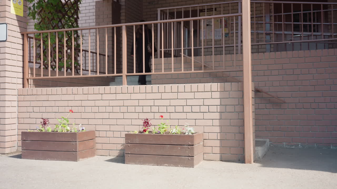 어두운 코트를 입은 학생이 벽돌 계단을 올라 측면 입구로 향하고, 난간, 벤치, 쓰레기통이 있는 조용한 안뜰을 가로지르며 긴 그림자가 드리워진 한낮의 햇살 아래 경사로 옆 문으로 꾸준히 다가가고 있어요.