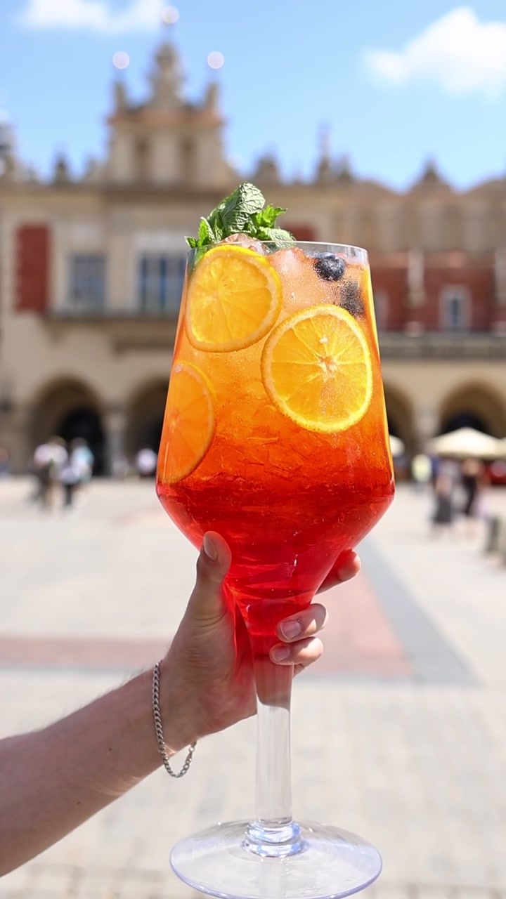 Mão Segurando Grande Coquetel Aperol Spritz em Praça de Cidade Europeia
