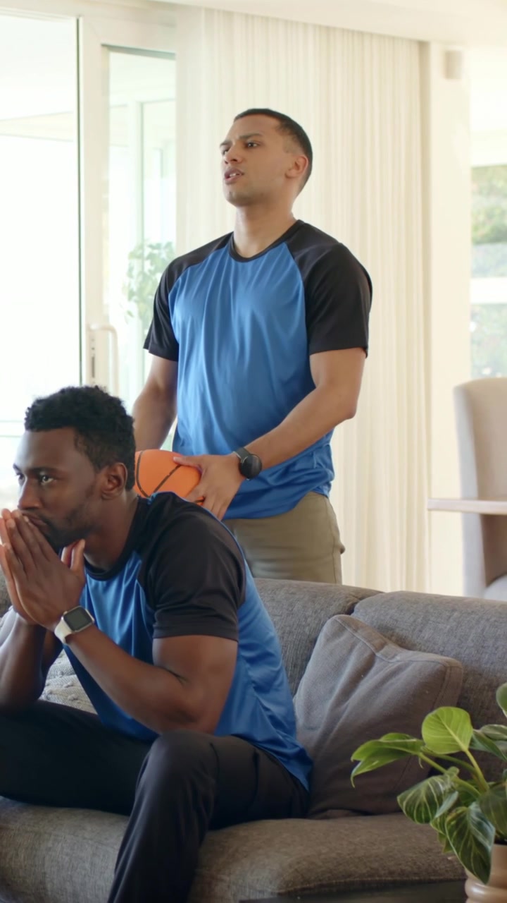Vertical video: Watching off-camera play, duo in blue shirts gesturing on sofa with basketball