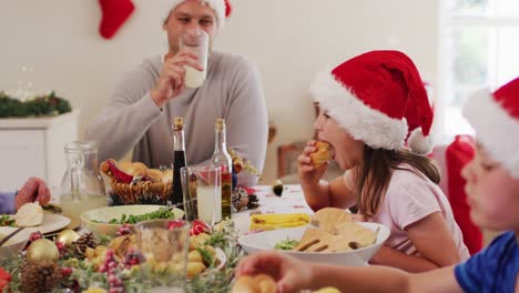 Kaukasische-Familie-In-Weihnachtsmützen-Genießt-Gemeinsam-Das-Mittagessen,-Während-Sie-Zu-Hause-Am-Esstisch-Sitzt